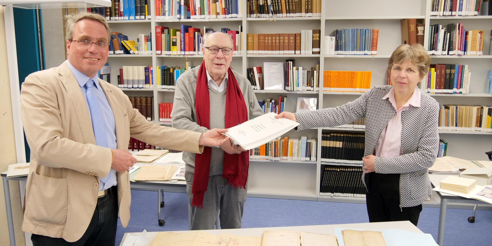 Übergabe im Diözesanarchiv: Dr. Peter Nieveler (M.) und Pfarrer Josef Wolff (l.) nehmen das Findbuch von Dr. Beate Sophie Fleck, Leiterin des Diözesanarchivs Aachen, entgegen. Geschichte zum Anfassen: Viele Pfarrarchive im Bistum Aachen reichen oft zurück bis ins Mittelalter. Sie erzählen nicht nur die Historie der Kirche, sondern auch die Entwicklung von Dörfern und Städten, von Kriegen und Hungersnöten. Aufgrund der Fülle von Handschriften, Kirchenbüchern und Akten wird es für die Gemeinden immer schwieriger geeignete Räume zu finden, um die wertvollen Dokumente sowohl aus konservatorischen Gesichtspunkten als auch aus dem Blickwinkel des Datenschutzes heraus sicher unterzubringen. Immer mehr Gemeinden nutzen deswegen die Möglichkeit, ihre Pfarrarchive als Depositum dem Bischöflichen Diözesanarchiv des Bistums Aachen zu übergeben – so auch jüngst die Pfarrei Heilig Geist in Jülich. Sie übergab das Pfarrarchiv der ehemaligen Pfarrei Selgersdorf St. Stephanus, die seit 2013 als Gemeinde zur Pfarrei Heilig Geist Jülich gehört, an das Diözesanarchiv.