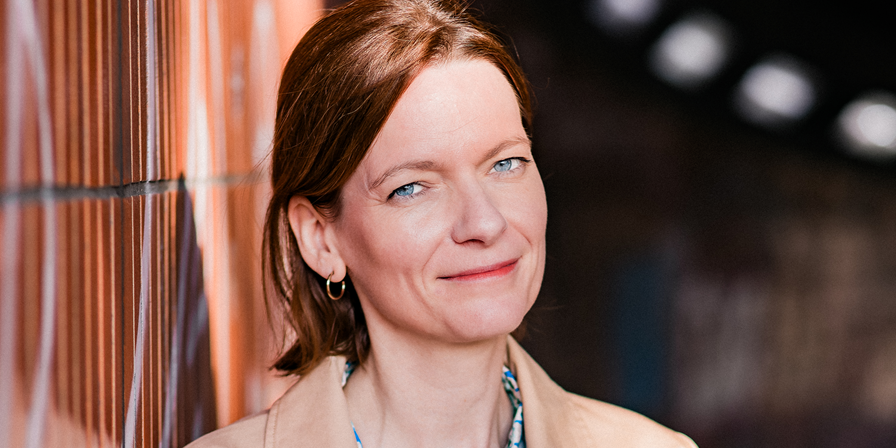 Frau, helle Bluse, braune Haare, an Mauer gelehnt