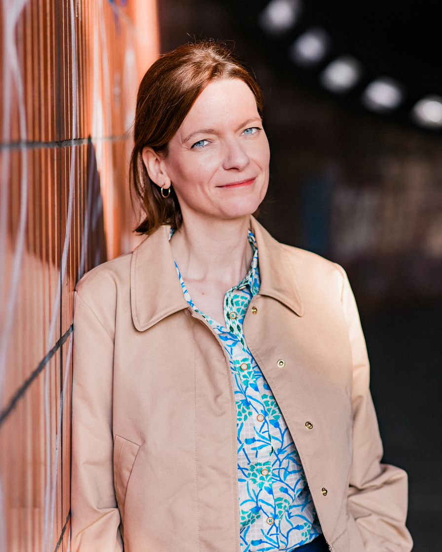 Frau, helle Bluse, braune Haare, an Mauer gelehnt