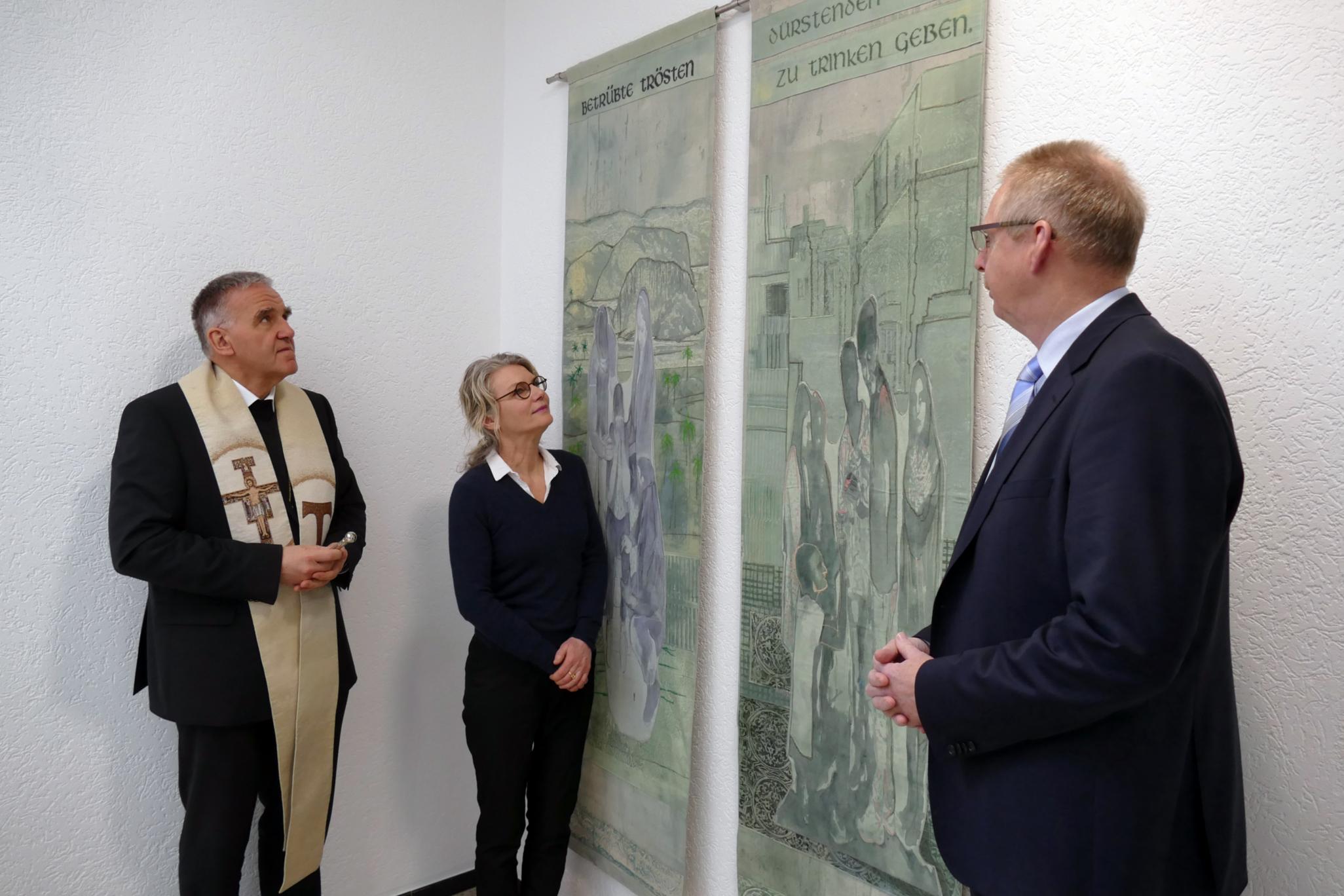 Der Vorsitzende des Caritasverbandes für das Bistum Aachen, Weihbischof Dr. Johannes Bündgens (.), und die Künstlerin Vera Sous erläuterten die Banner und ihren Entstehungsprozess. Diözesancaritasdirektor Burkard Schröders (r.) freut sich, dass die Kunstwerke nun im Haus der Caritas hängen.