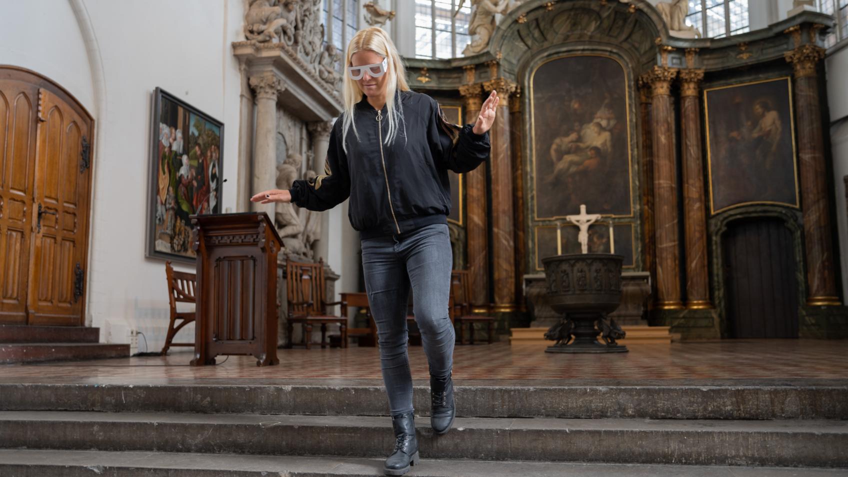 Foto: Eine junge Frau mit einer großen Brille aus weißer Pappe steigt vorsichtig Treppenstufen hinunter, mit ausgebreiteten Armen hält sie das Gleichgewicht. Hinter ihr der Altarraum einer Kirche. Sie hat lange, blonde Haare und trägt schwarze Kleidung. (c) DBSV-Friese