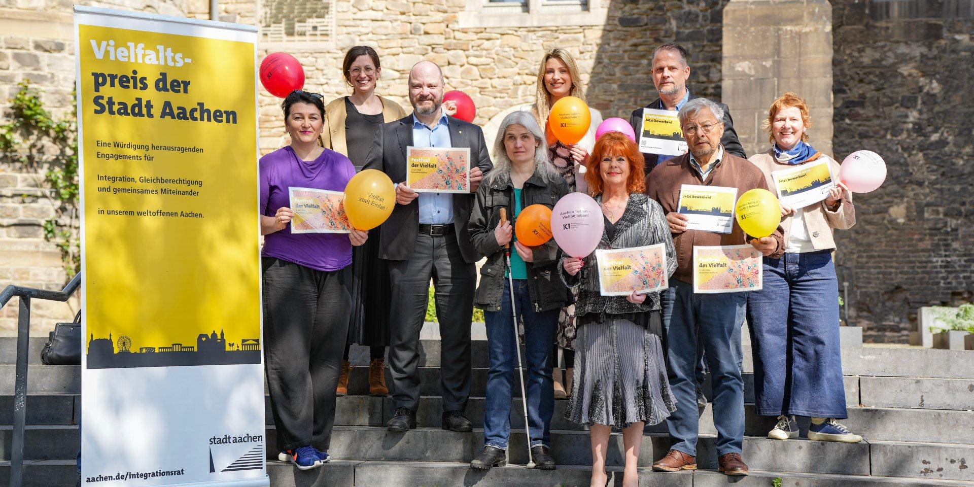 Unterschiedliche Meschen stehen als Gruppe auf einer Treppe und halten bunte Luftballons
