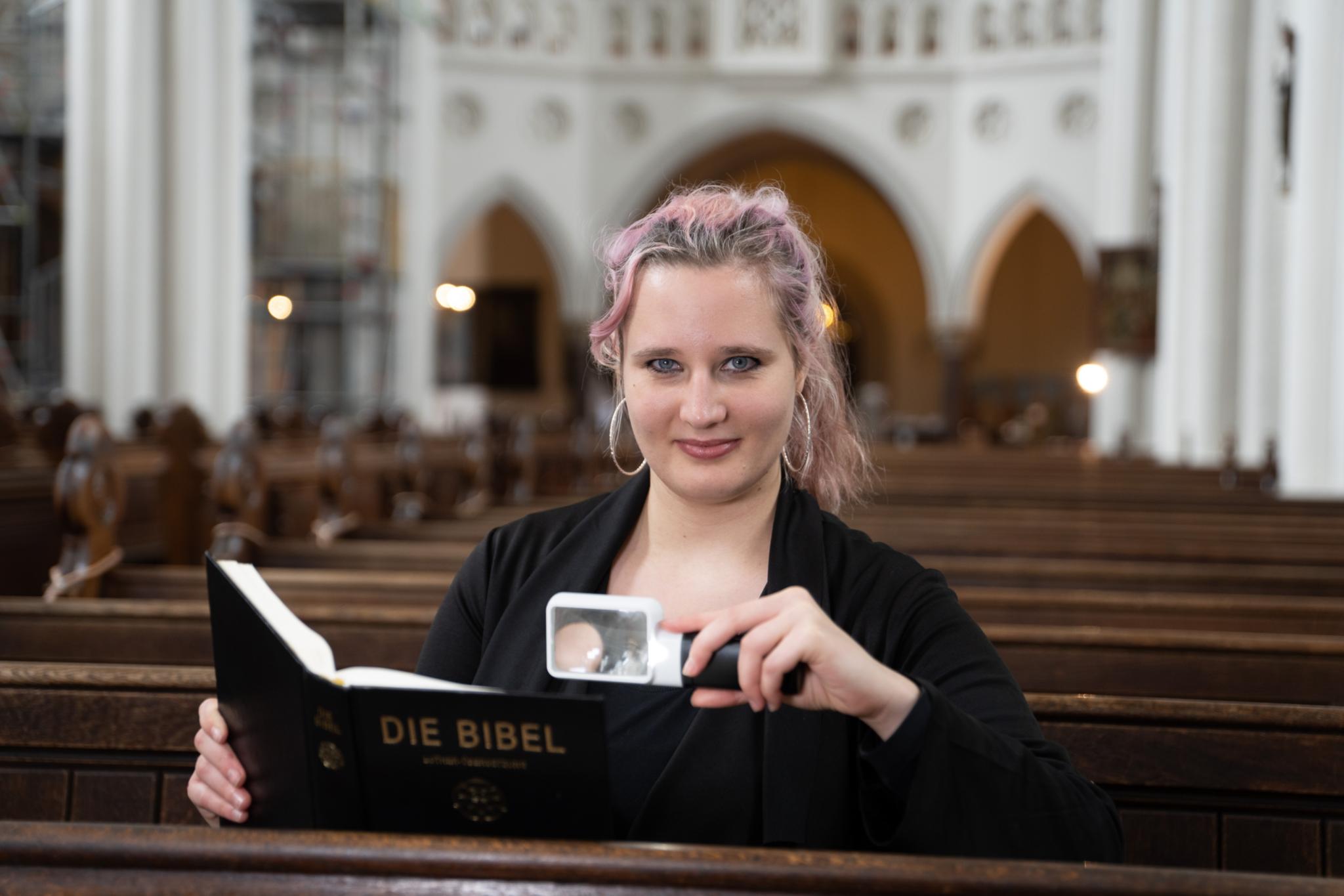 Foto: Eine Frau Mitte 20 sitzt in einer Kirchenbank und lächelt in die Kamera, in einer Hand eine Leuchtlupe, in der anderen eine Bibel. Sie hat langes, dunkelblondes Haar mit rötlichen Strähnchen und trägt große Ohrringe.