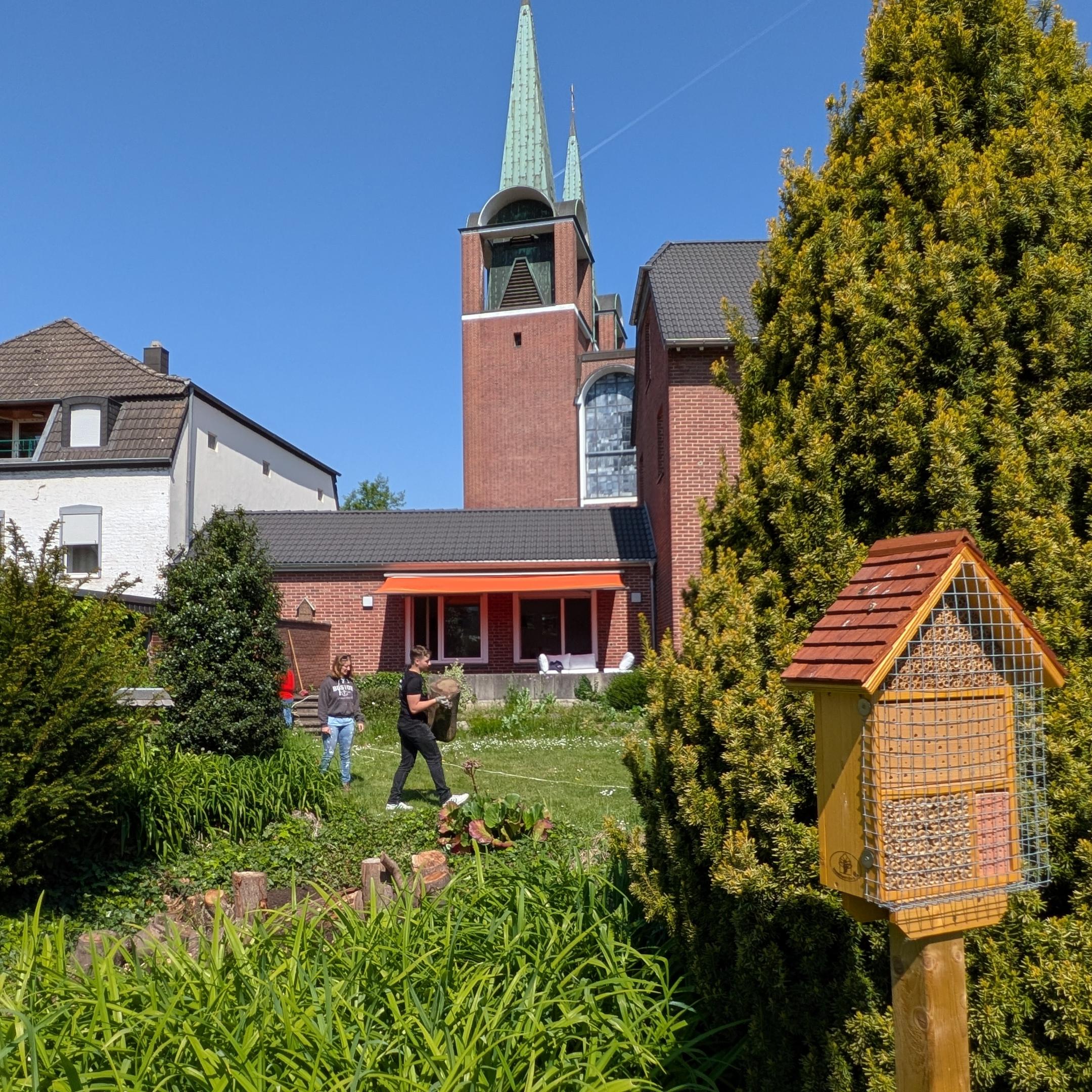 Das Bild zeigt einen Garten mit viel Wiese und einem Vogelhaus. Im Hintergrund sind ein Haus und eine Kirche