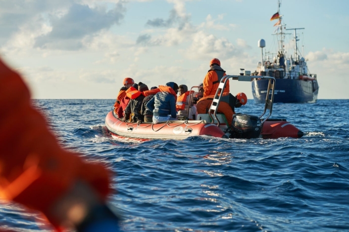 Szene aus dem Dokumentarfilm 'Route 4'. Geflüchtete auf einem zu kleinen Boot auf dem Meer, im Hintergrund ein Schiff.