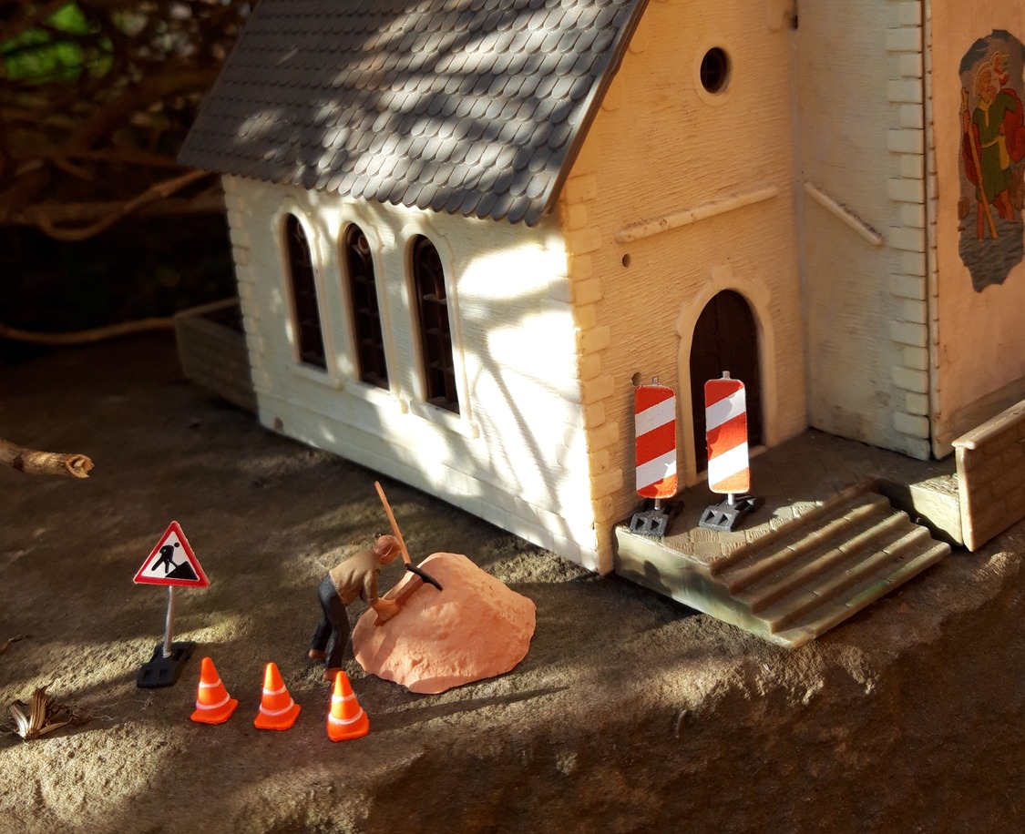 Vor einem Kirchengebäude im leichten Sonnenlicht ist eine Baustelle eingerichtet, ein Bauarbeiter arbeitet an einem Hügel mit Sand.