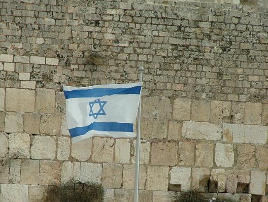 Israelische Flagge (zwei Streifen mit Davidsstern) vor einer Steinmauer.