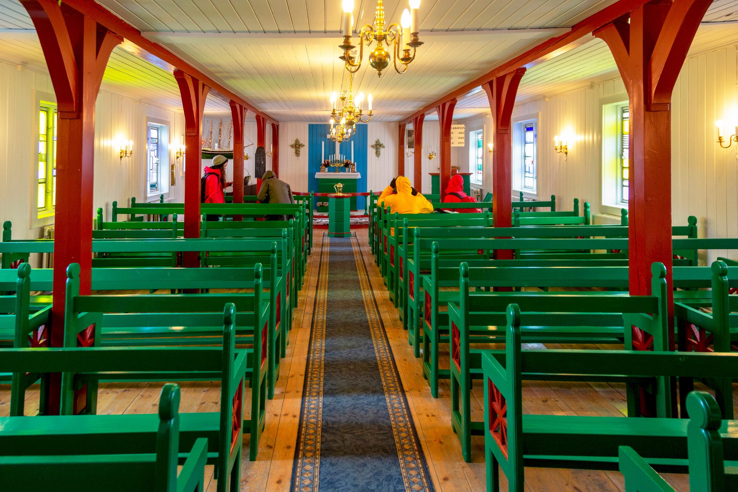 Das Innere einer lutherischen Kirche in Kulusuk auf Grönland, vornehmlich aus Holz gebaut, mit Bänken rechts und links des Mittelgangs. Seitlich sind Bleiglasfenster der Aachener Glasmalerin Maria Katzgrau zu sehen.