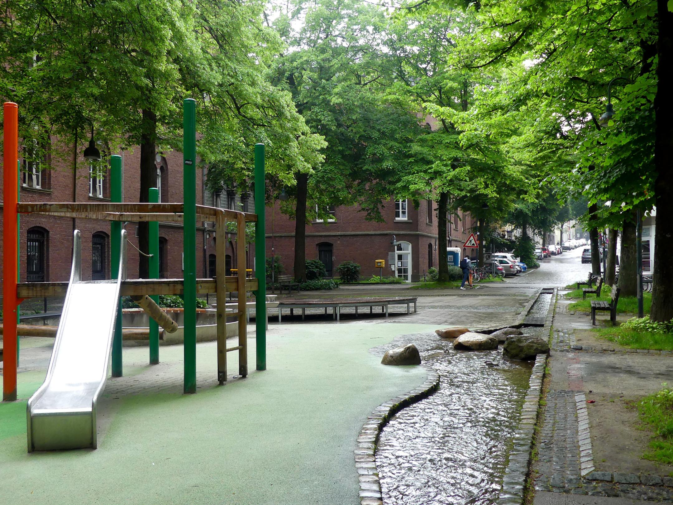 Ein Platz in der Innenstadt mit Kinderrutschbahn, ein schmaler Wasserlauf im Boden.