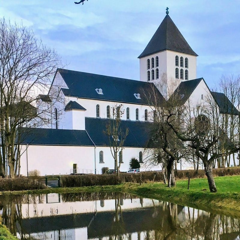 Ein Klostergebäude mit Kirchenschiff und Kirchturm inmitten eines Gartens mit einem kleinen Weiher davor.