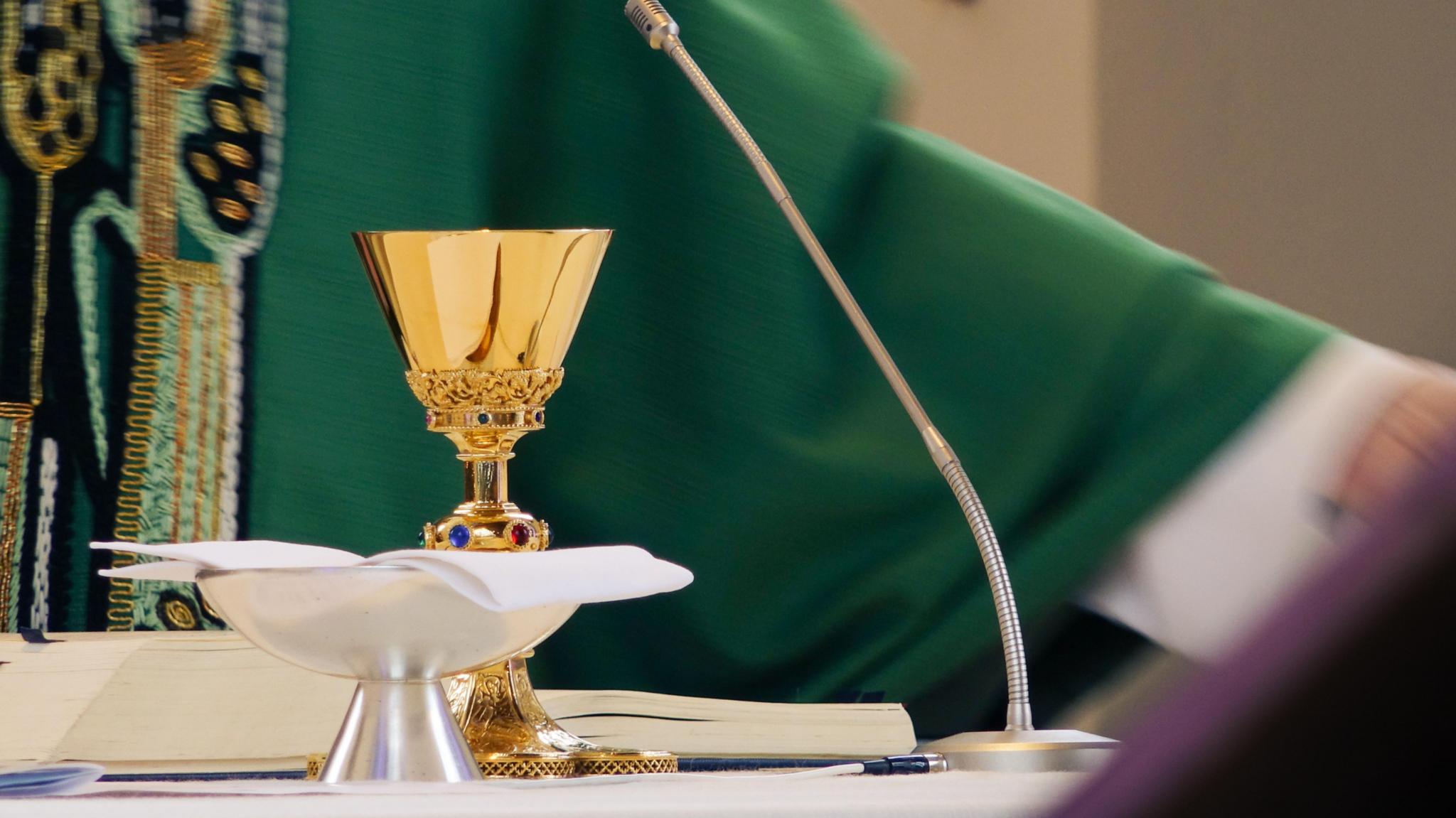Auf dem Altar stehen Kelch und Patene, dahinter sieht man einen Teil eines Priesters im grünen Messgewand.
