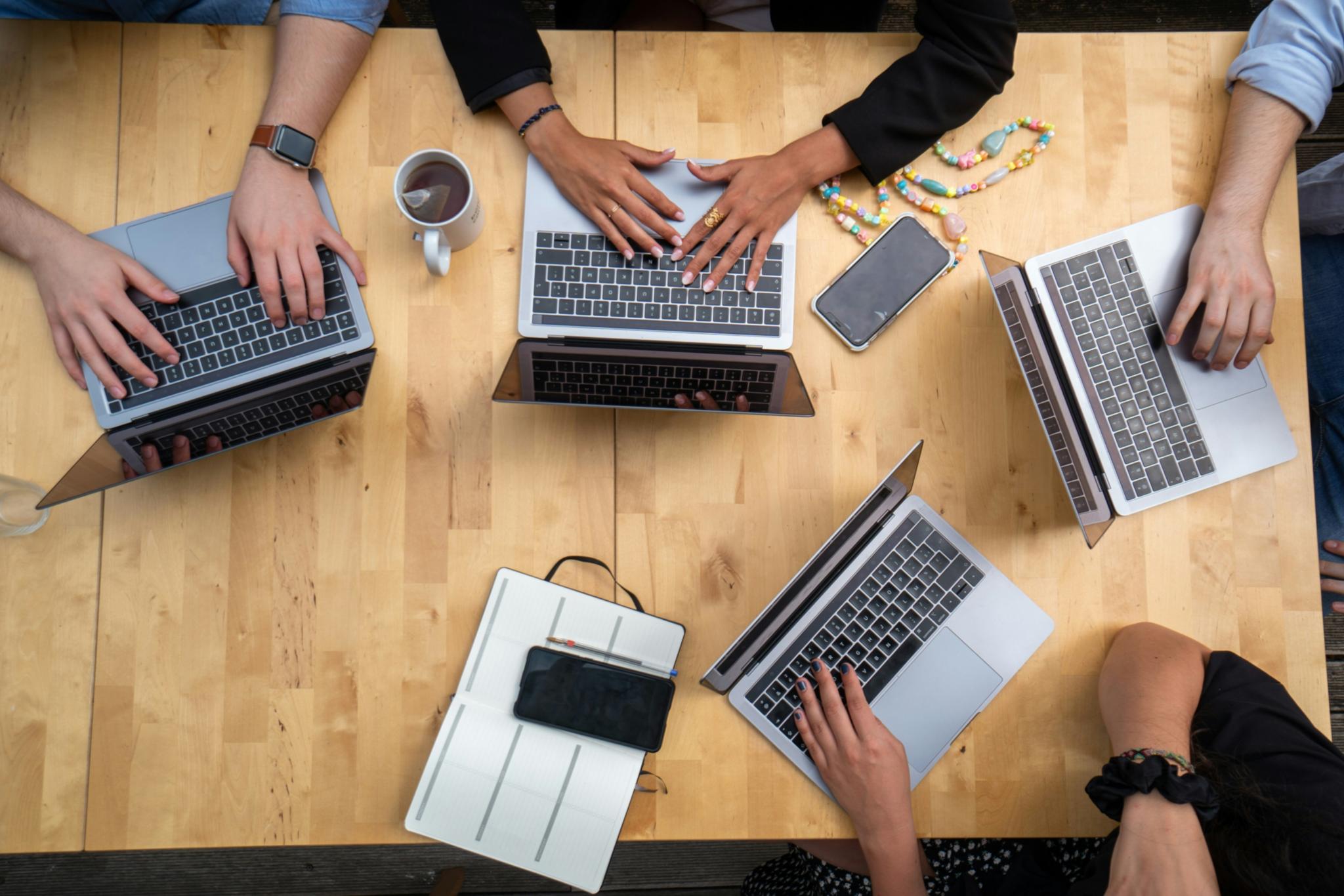 Blick auf Tischplatte, Menschen haben darauf ihre Laptops aufgeklappt und die Hände auf der jeweiligen Tastatur. Da die Hände unterschiedliche Hautfarben haben, dient das Bild zur Illustration des Themas 'Rassismus begegnen' im beruflichen Alltag.