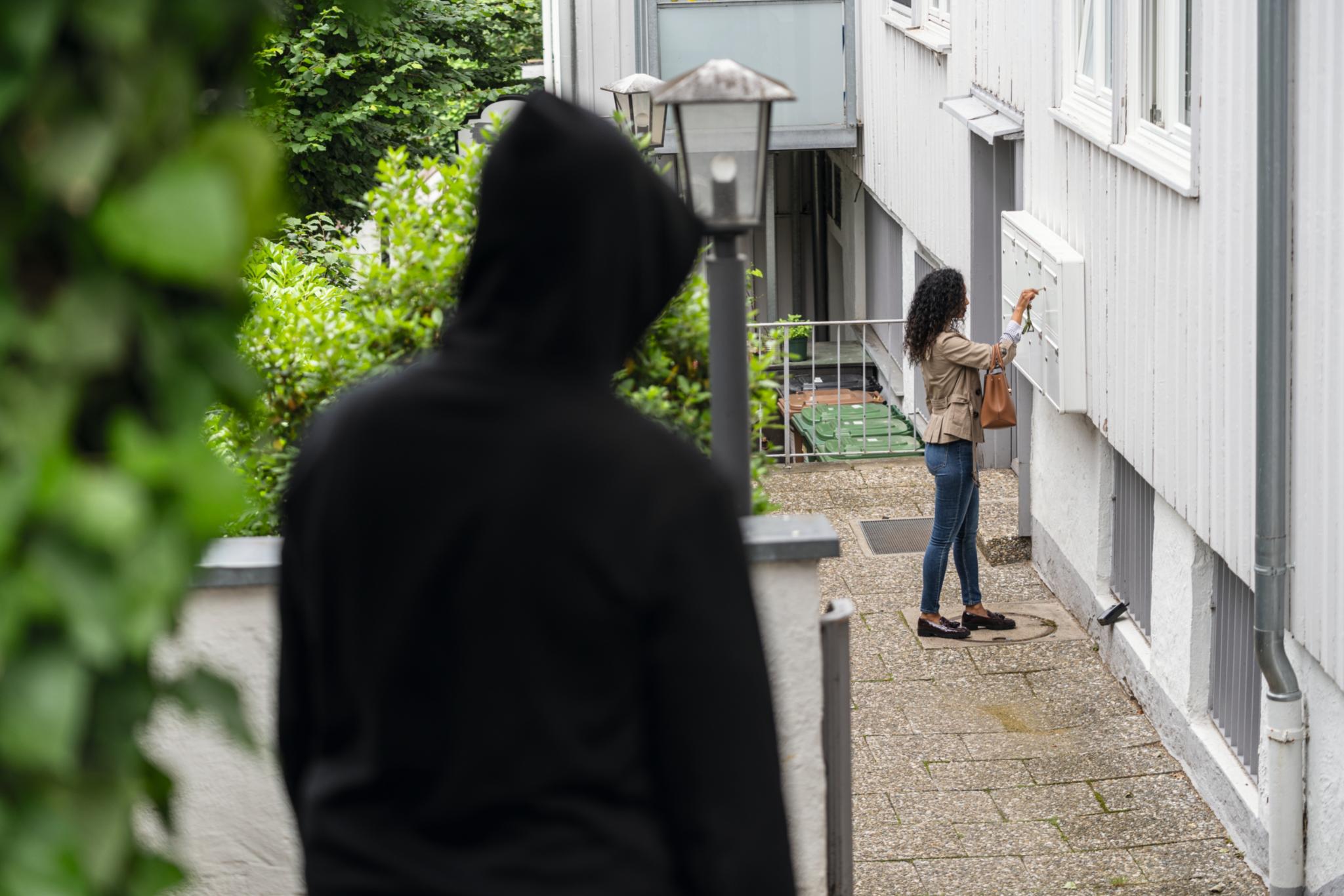 Frau steht an ihrer Wohnungstür und öffnet dort den Briefkasten, während ein Mann sie beobachtet