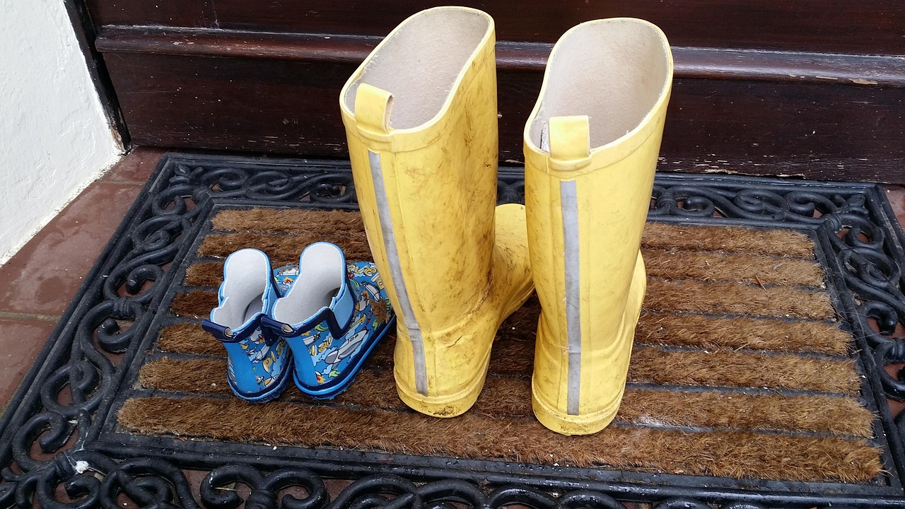 Nebeneinander auf einer Fußmatte steht ein Paar Gummistiefel eines Kindes (blau) neben einem Paar für Erwachsene (gelb).