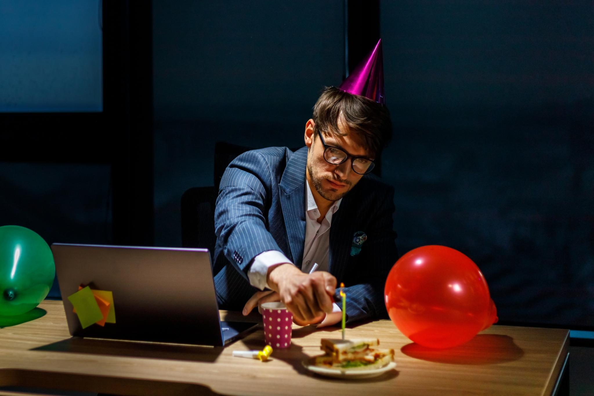 Ein Chef sitzt mit Partyhut alleine vor dem Laptop, neben ihm Luftballons, er zündet eine Kerze auf einem Geburtstagskuchen an.