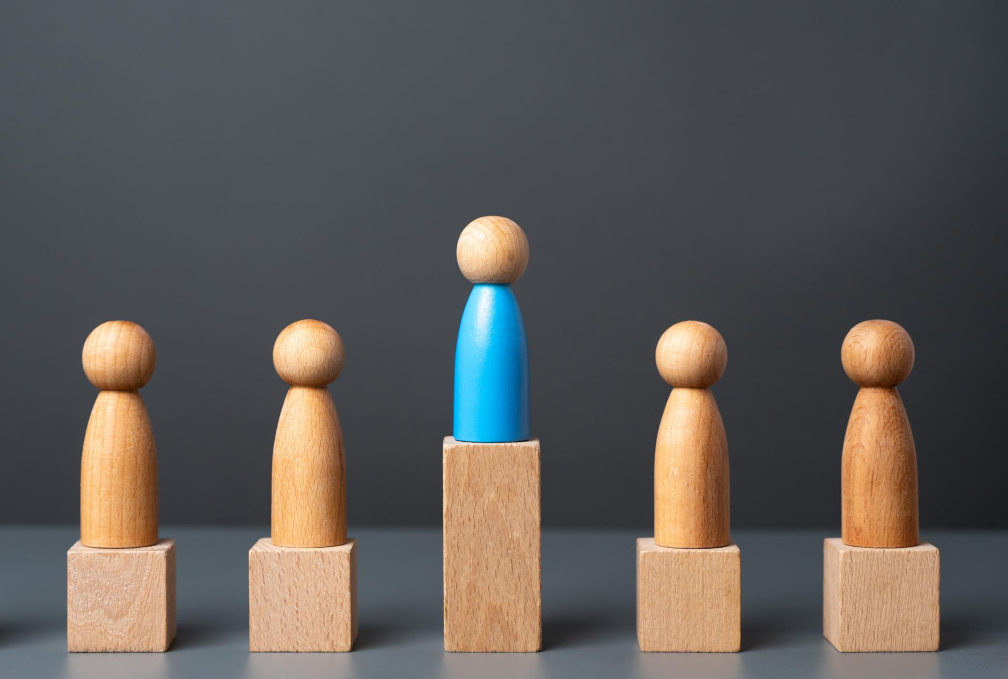 Fünf Holzspielfiguren auf kleinen Holzpodesten, die mittlere Figur steht auf einem höheren Podest, sie überragt die anderen.