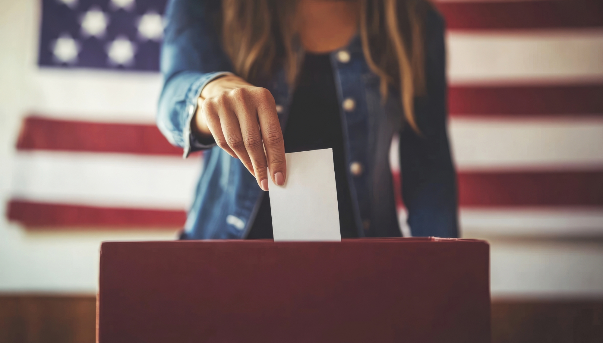 Eine Frau steckt einen Wahlzettel in eine Wahlurne, das Gesicht ist nicht erkennbar. Hinter ihr die Flagge der USA.