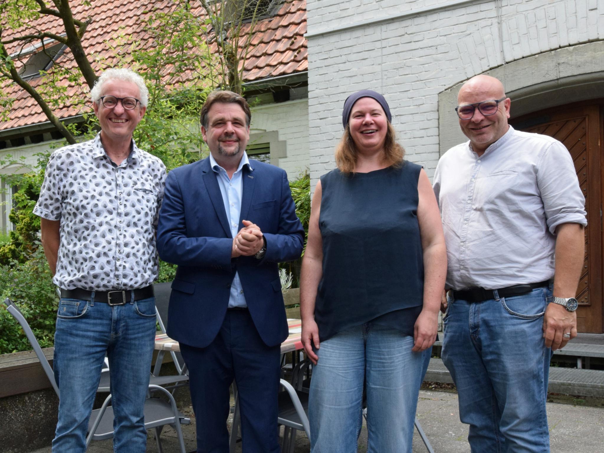 Drei Männer und eine Frau stehen in einem Innenhof vor einem Tor und einem Baum.