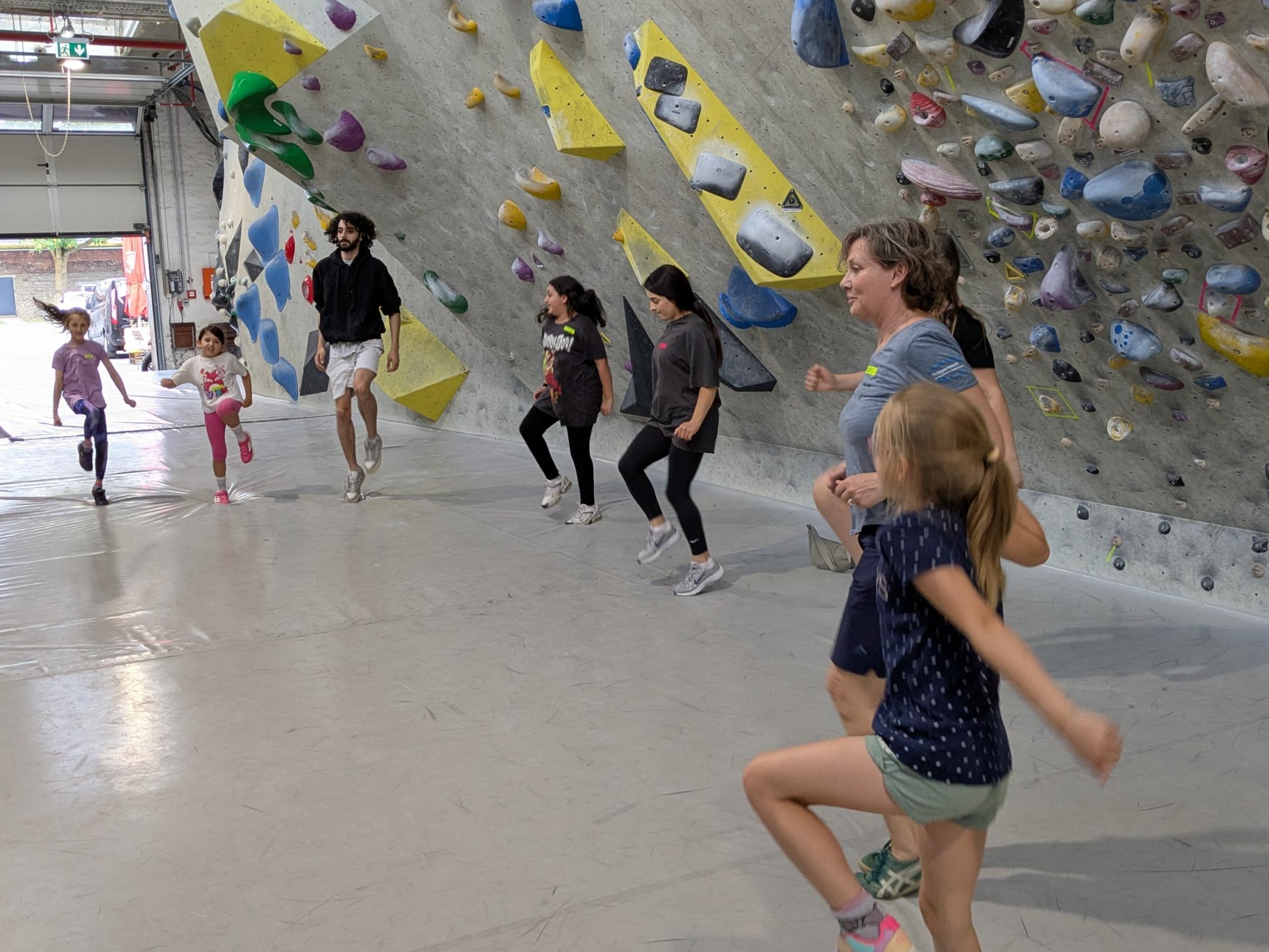 Kinder springen im Halbkreis als Aufwärmübung vor einer Kletterwand.