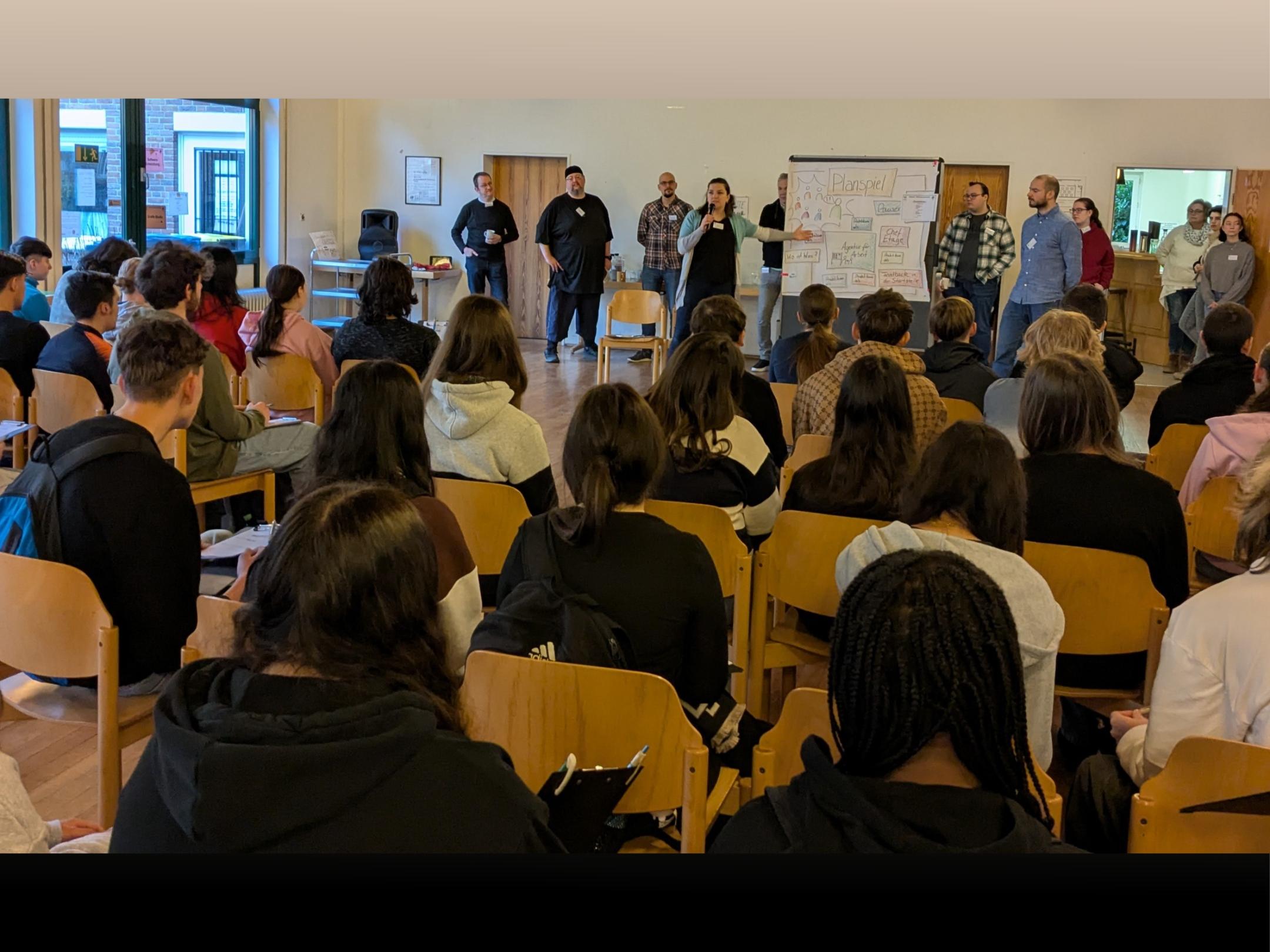 Viele Leute sitzen wie in einer Klasse und schauen nach vorne auf eine Tafel, wo einer etwas erklärt. Daneben stehen weitere Leute.