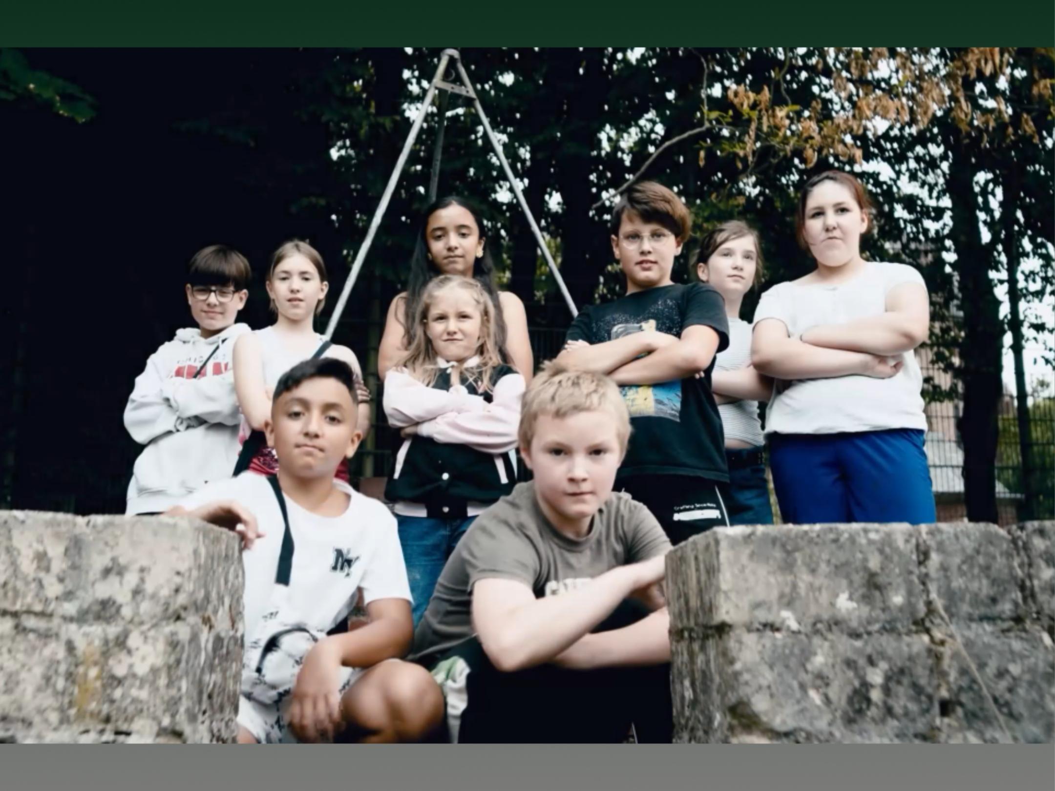 9 Kinder stehen, bzw. hocken hinter einer kleinen Mauer mit Öffnung und verschränken teilweise die Arme.