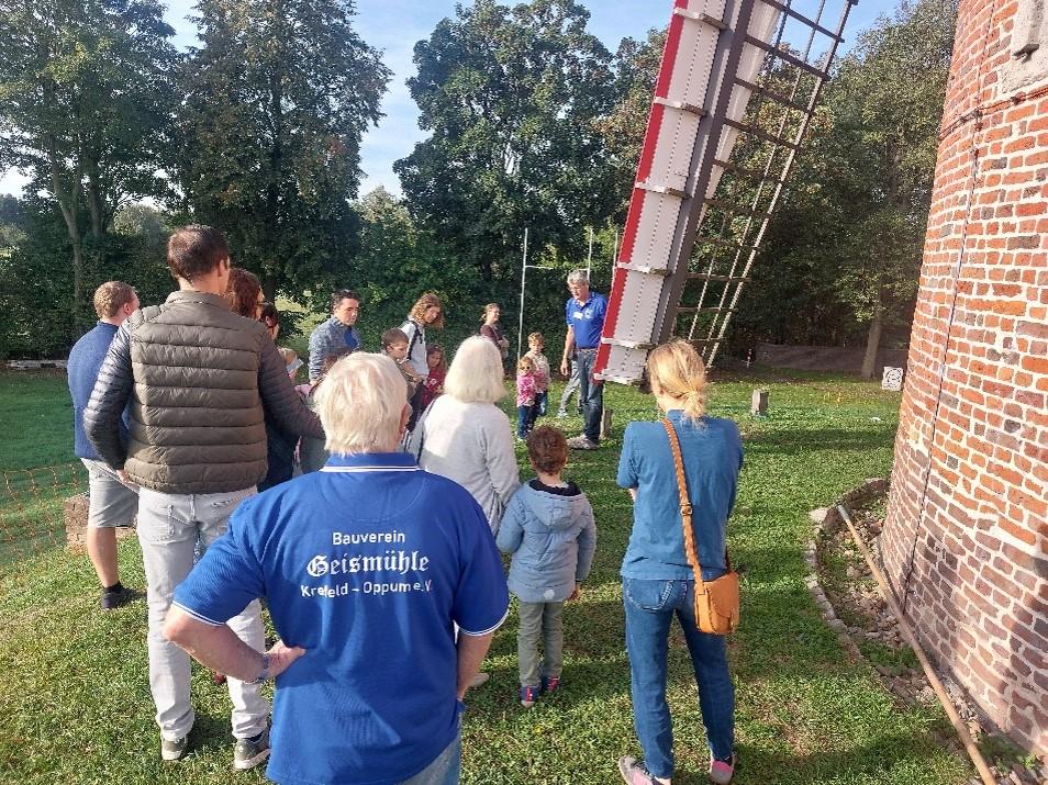Mehrere Personen stehen mit dem Rücken zum Fotografen, Rechts sind Teile der Mühle erkennbar (Backsteine und ein Teil des Windfügels), im Hintergrund Bäume und grüne Wiese.